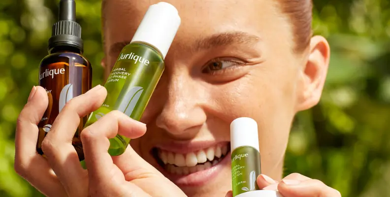 Smiling person holding Jurlique skincare bottles, including a serum and facial oil, against a green, natural background.