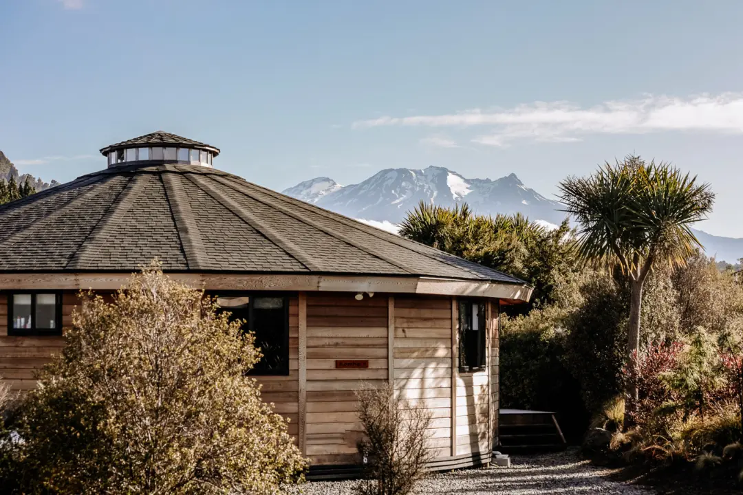 Round wooden cabin with a shingled roof, surrounded by trees, with snow-capped mountains in the background.
