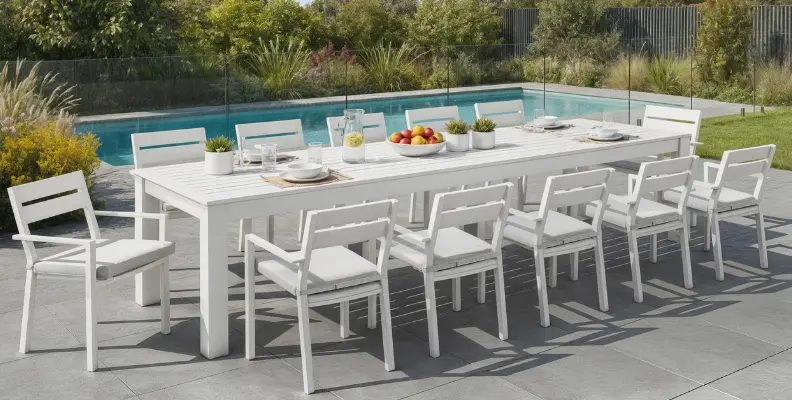 Outdoor dining setting with a long white table and ten matching chairs by a pool, featuring tableware and potted plants.