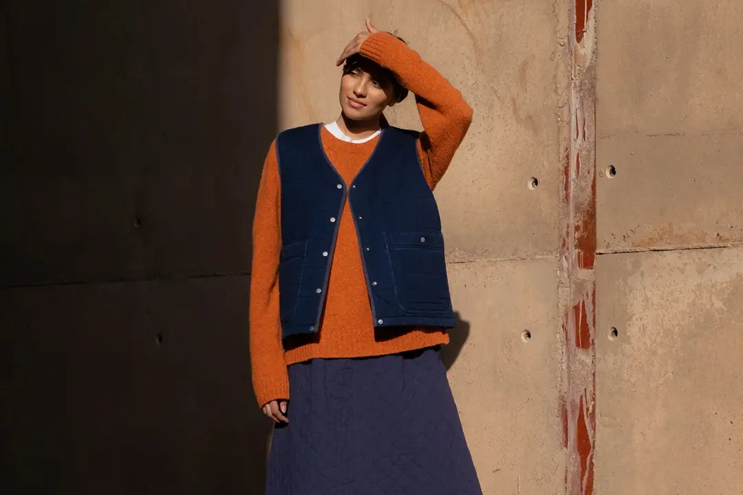 Person mit einem orangefarbenen Pullover, einer blauen Steppweste und einem marineblauen Rock, die vor einer Betonwand steht und ihre Augen vor der Sonne schützt.