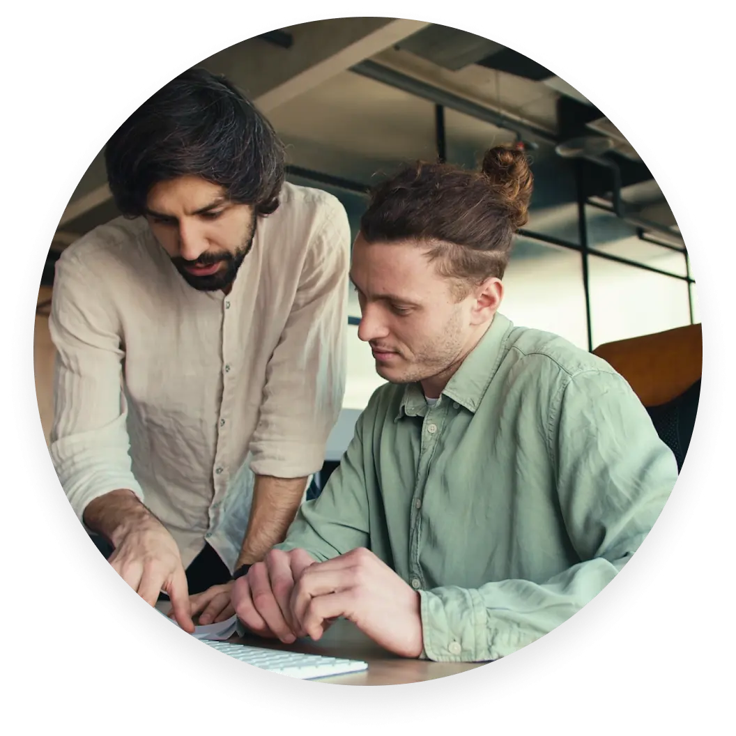 Dos personas colaborando en una mesa, una de pie y señalando un teclado y la otra sentada y concentrada