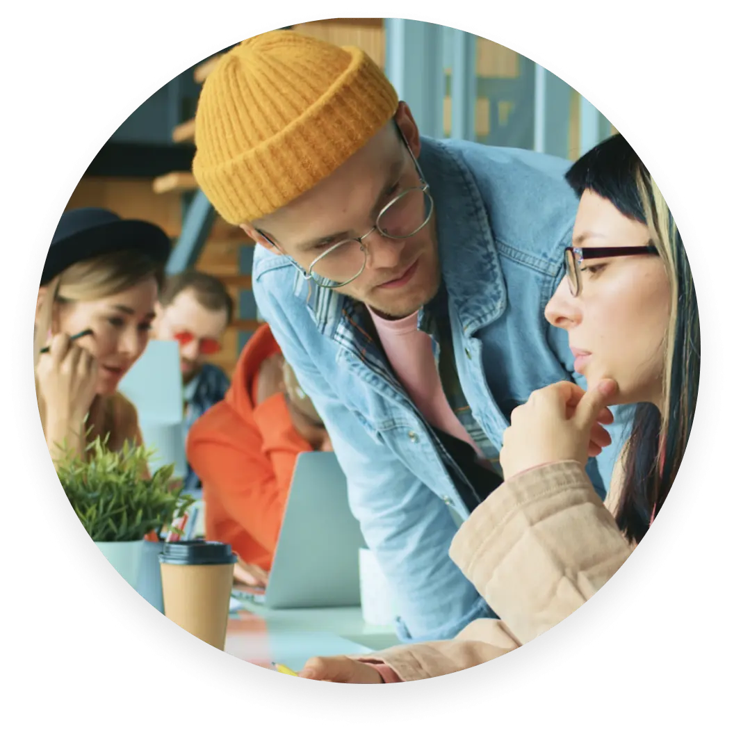 Dos personas en un entorno de oficina informal, una con un gorro amarillo y gafas inclinándose para hablar de algo con la otra