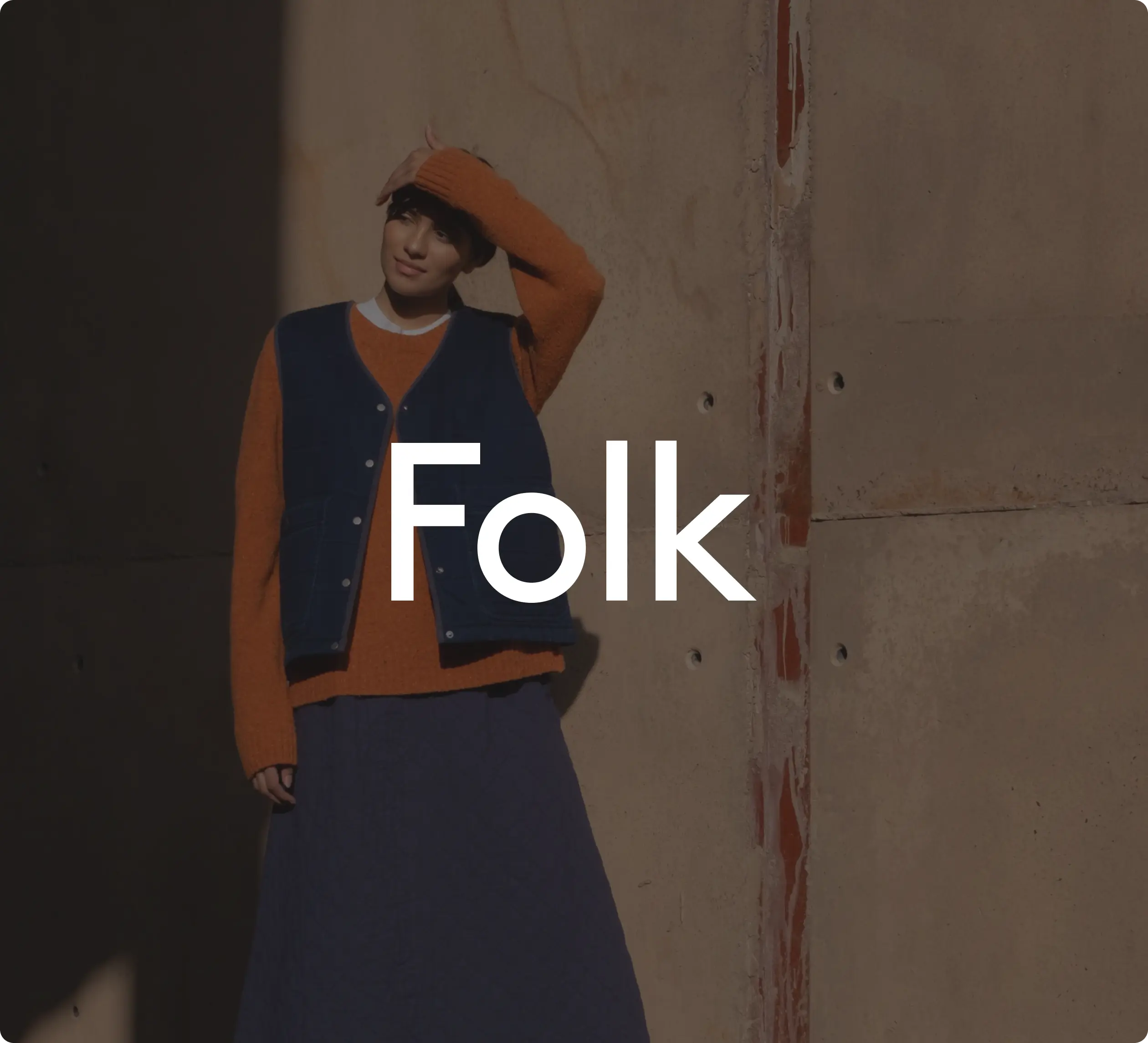 Personne portant un pull orange et un gilet bleu, se tenant contre un mur. Texte "Folk" au centre de l'image.