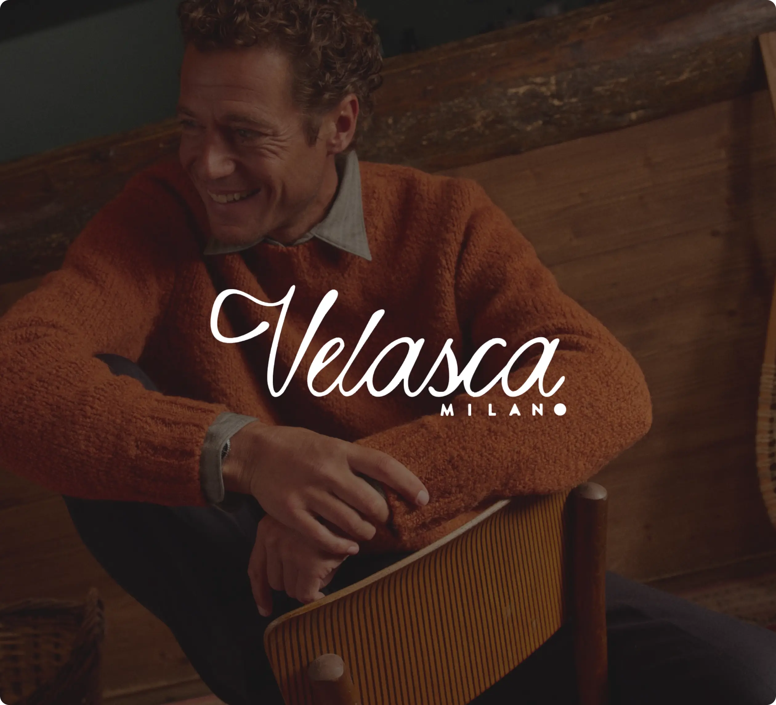 Homme souriant portant un pull orange, assis sur une chaise en bois. Logo "Velasca Milano" superposé.
