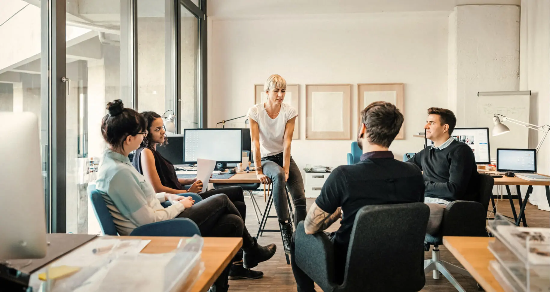 Groupe de cinq personnes en réunion dans un bureau moderne avec des ordinateurs et de grandes fenêtres