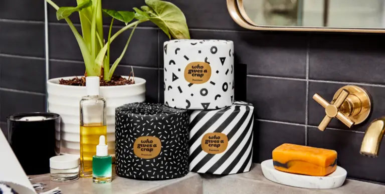 Alcuni rotoli di carta igienica con il marchio "Who Gives A Crap" posizionati in un bagno, insieme ad articoli da toeletta e una pianta in vaso.