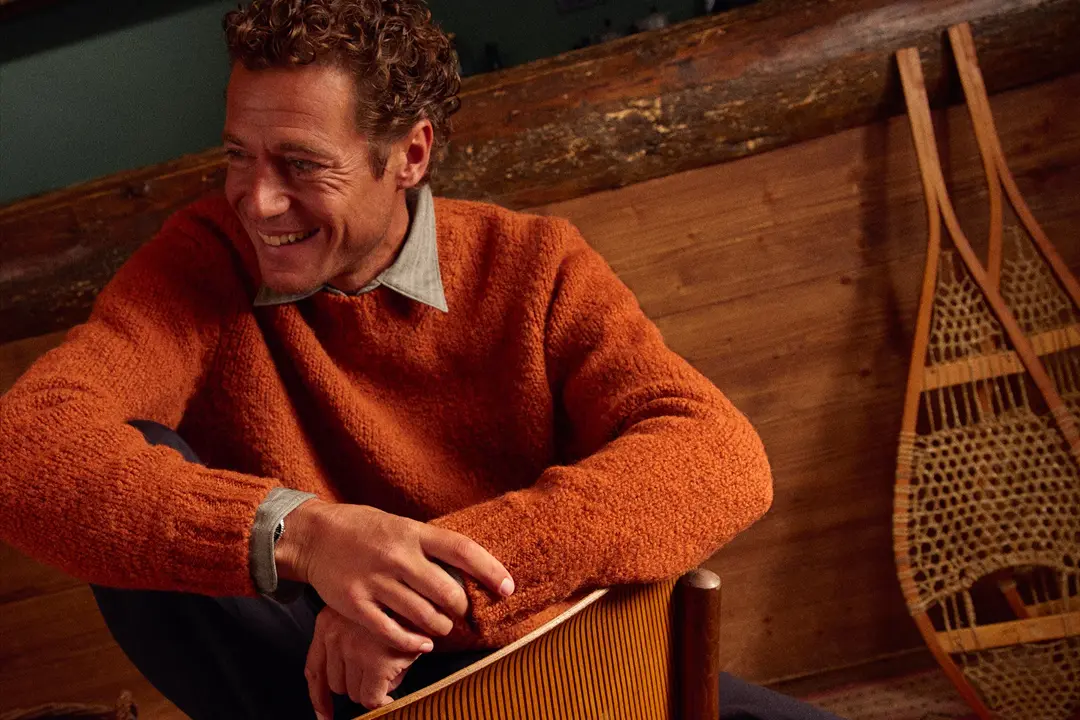 Un uomo sorridente indossa un maglione arancione ed è seduto su una sedia di legno. Sullo sfondo, delle racchette da neve.