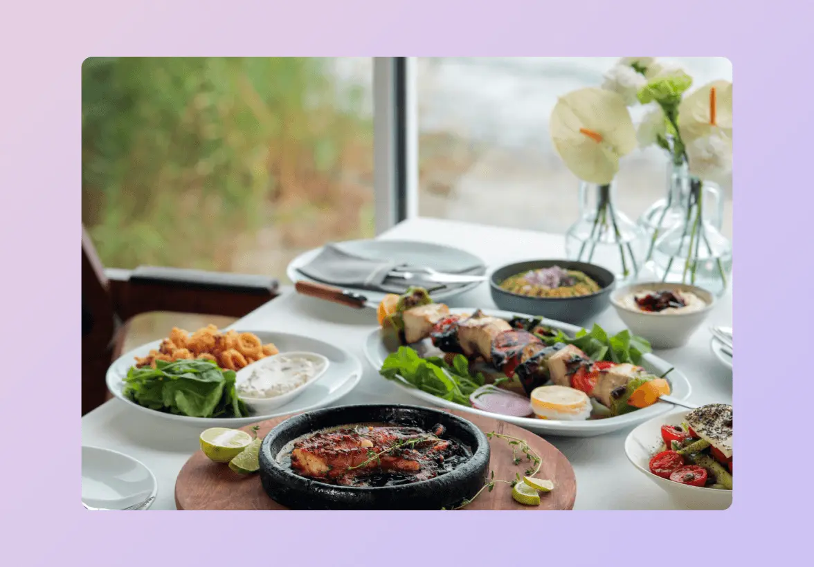 A table set with various dishes, including grilled meat, skewers, salad, and sauces, with flowers in vases in the background.