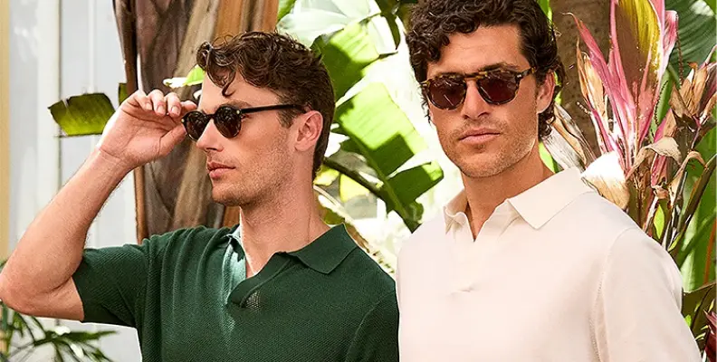Two men wearing sunglasses and casual shirts stand outdoors near tropical plants.