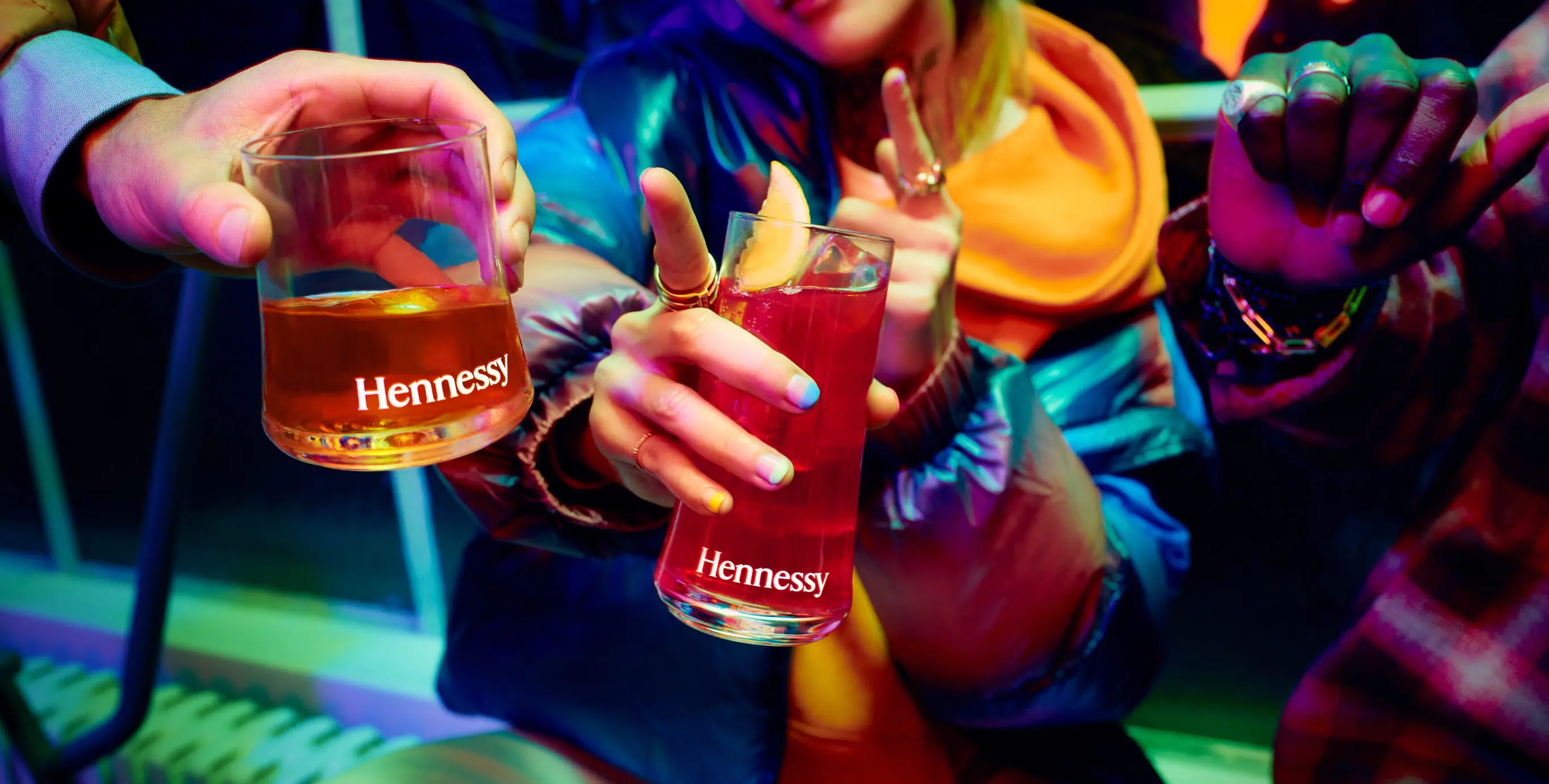 People holding Hennessy glasses in a vibrant setting, with colourful lighting and casual attire.