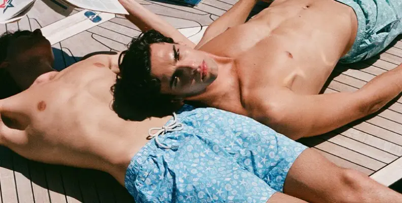 Two people sunbathing on a wooden deck, wearing patterned blue swim shorts.