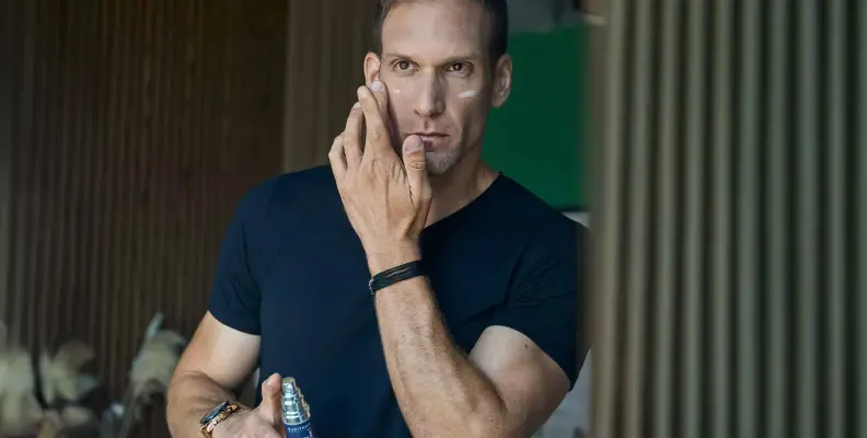 Man applying skincare product to face, holding a bottle in one hand, wearing a black T-shirt.