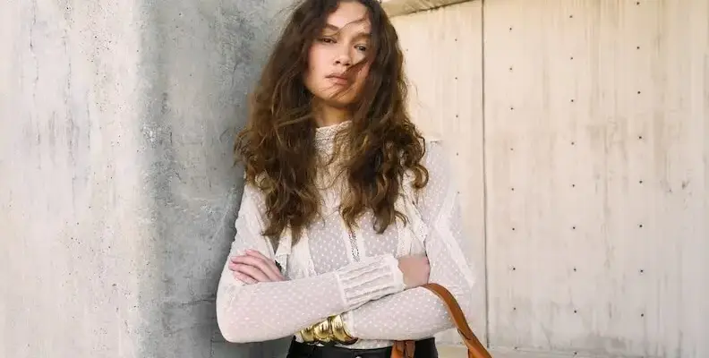 A woman with long, wavy hair wearing a white, textured blouse and gold bracelets stands against a concrete wall, holding a brown bag.