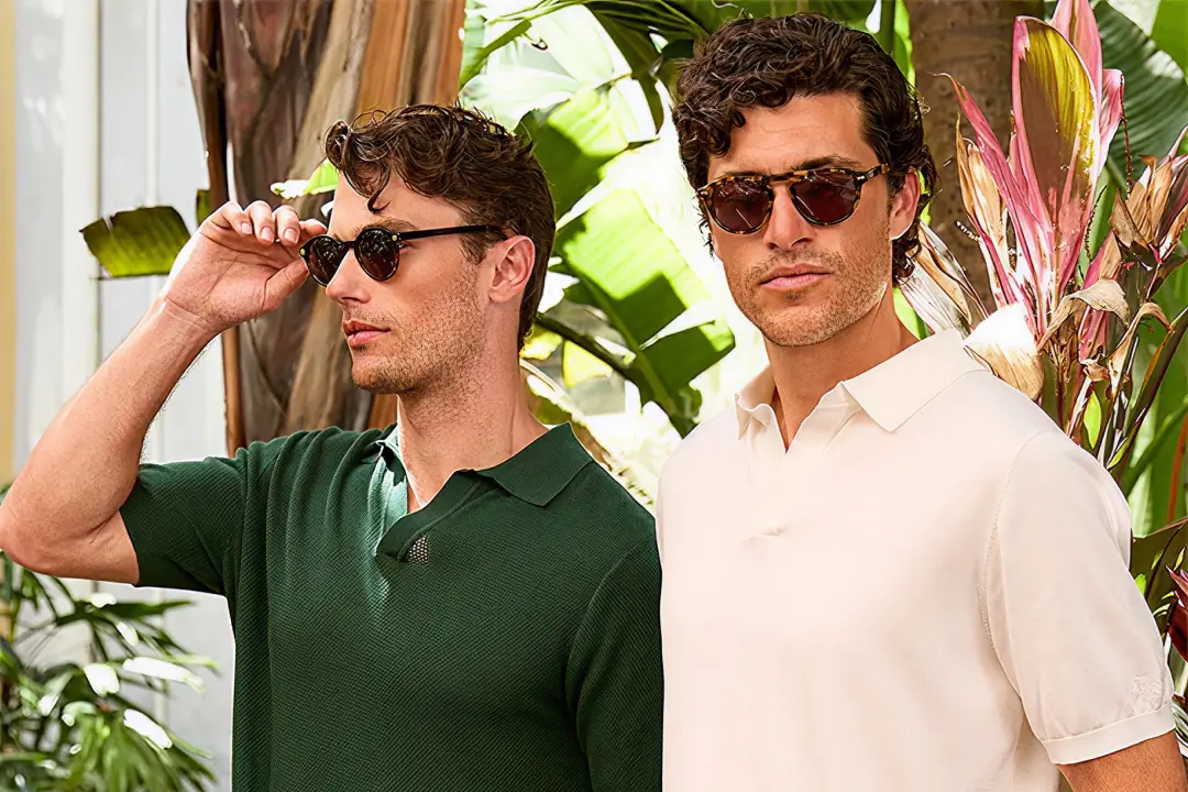 Two men wearing sunglasses, one in a green shirt and the other in a white shirt, standing outdoors with tropical plants.