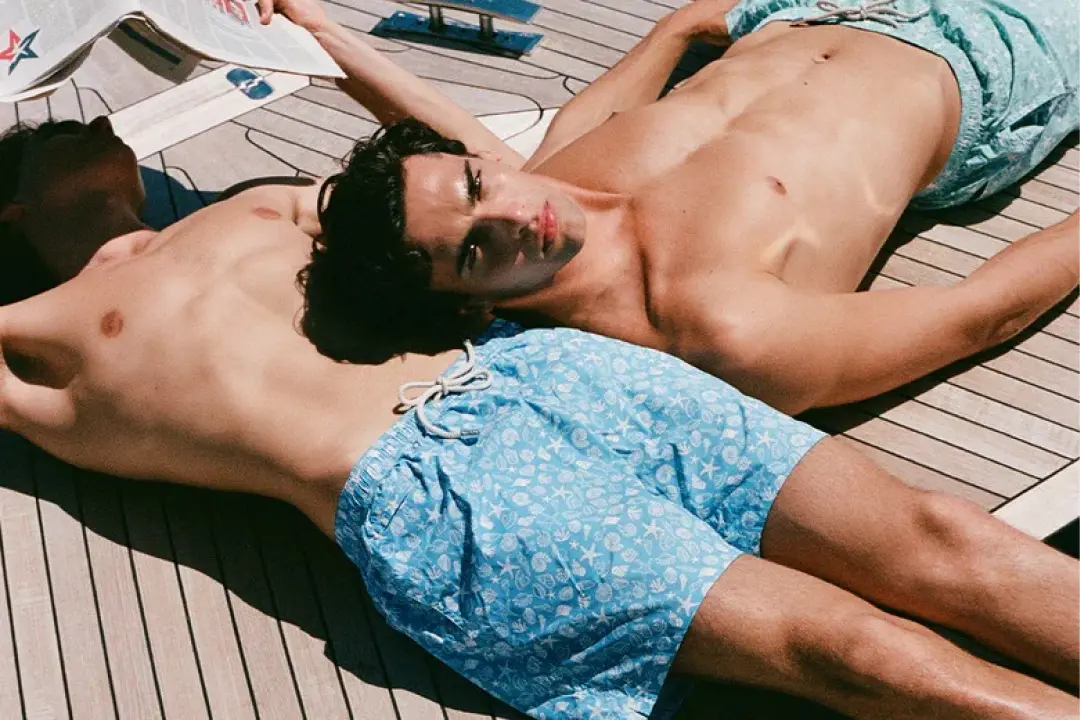 Two men sunbathing on a wooden deck, wearing patterned swim shorts. One holds a magazine.