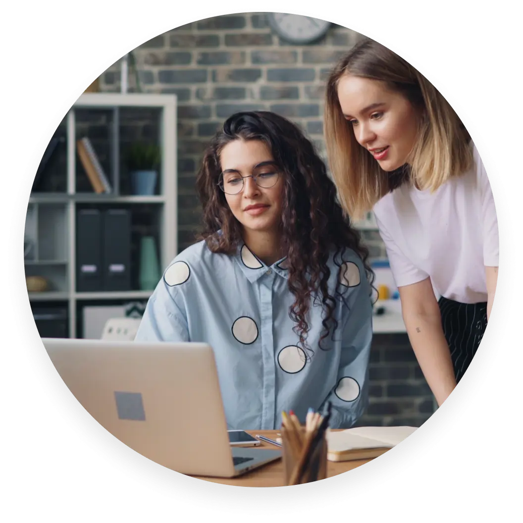 Two women looking at a laptop in an office setting, one seated and one standing, with shelves in the background.