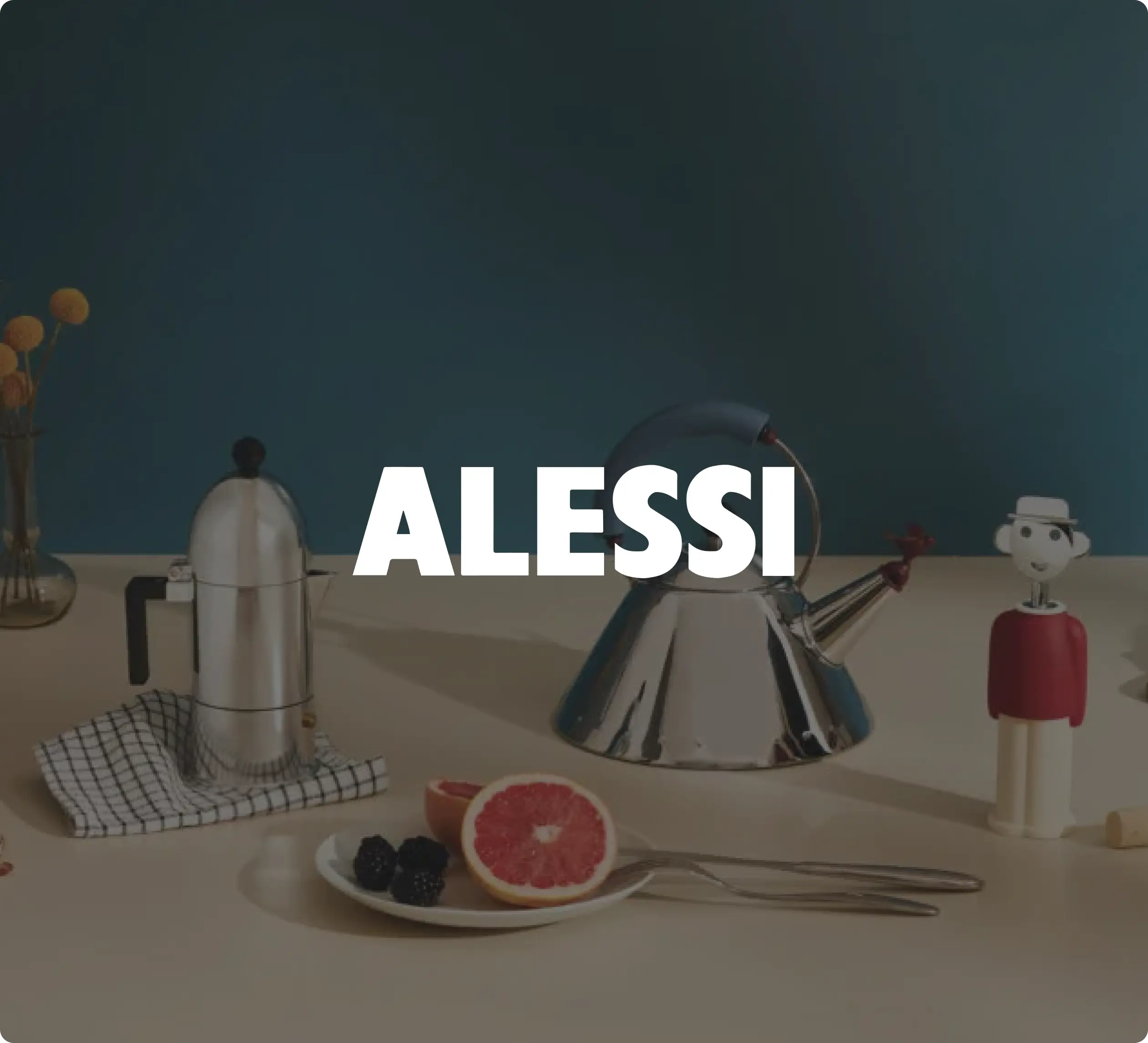 Alessi kitchenware on a table, including a kettle, coffee maker, and fruit plate. The word "ALESSI" is prominently displayed.