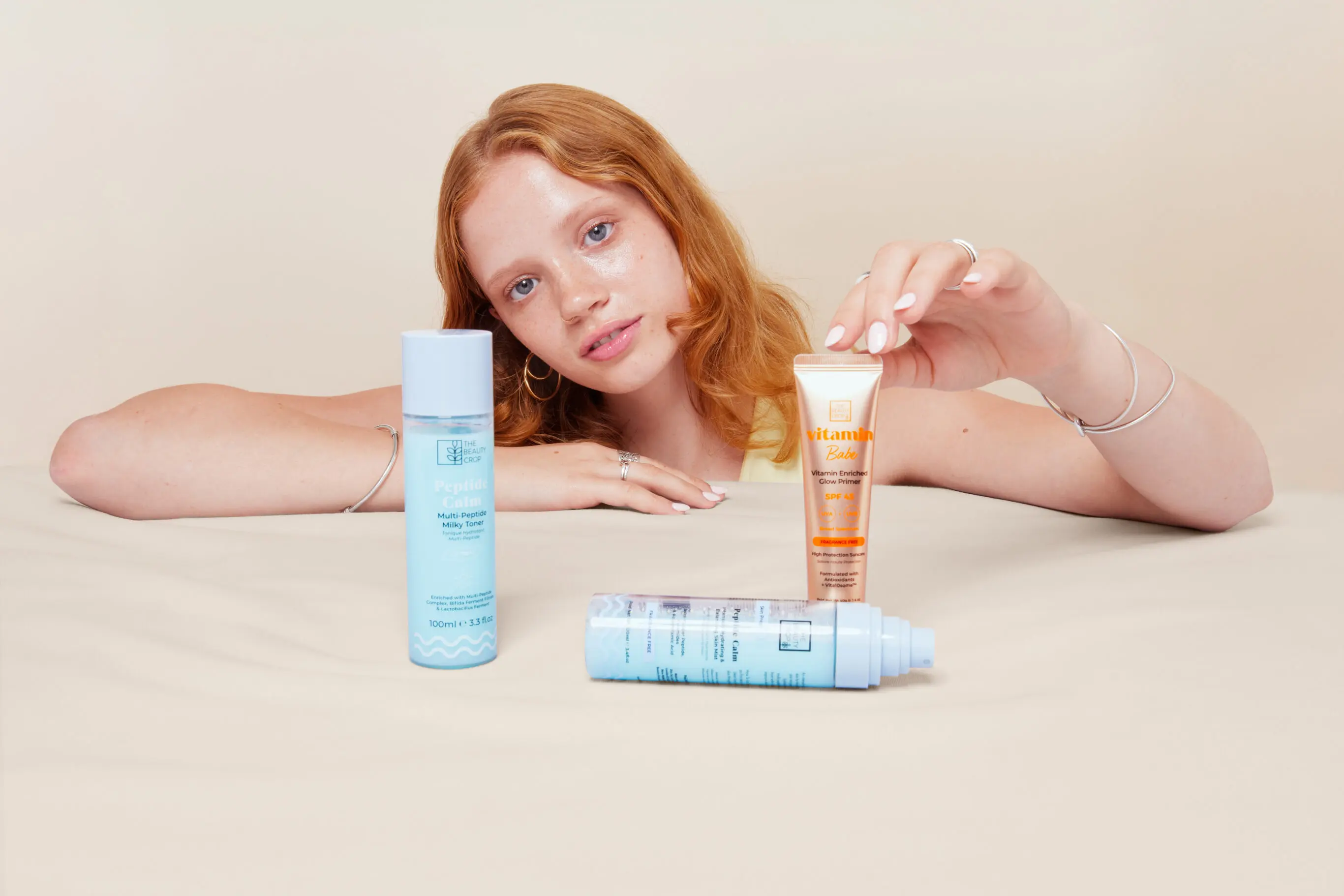 Woman with red hair showcasing The Beauty Crop skincare products, including Peptide Calm toner and Vitamin Babe primer.