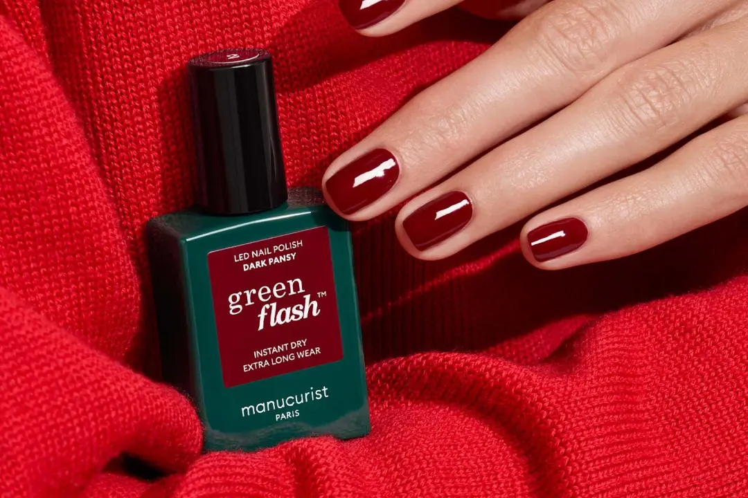 Red manicured nails next to a bottle of Manucurist Green Flash LED nail polish on a red fabric.