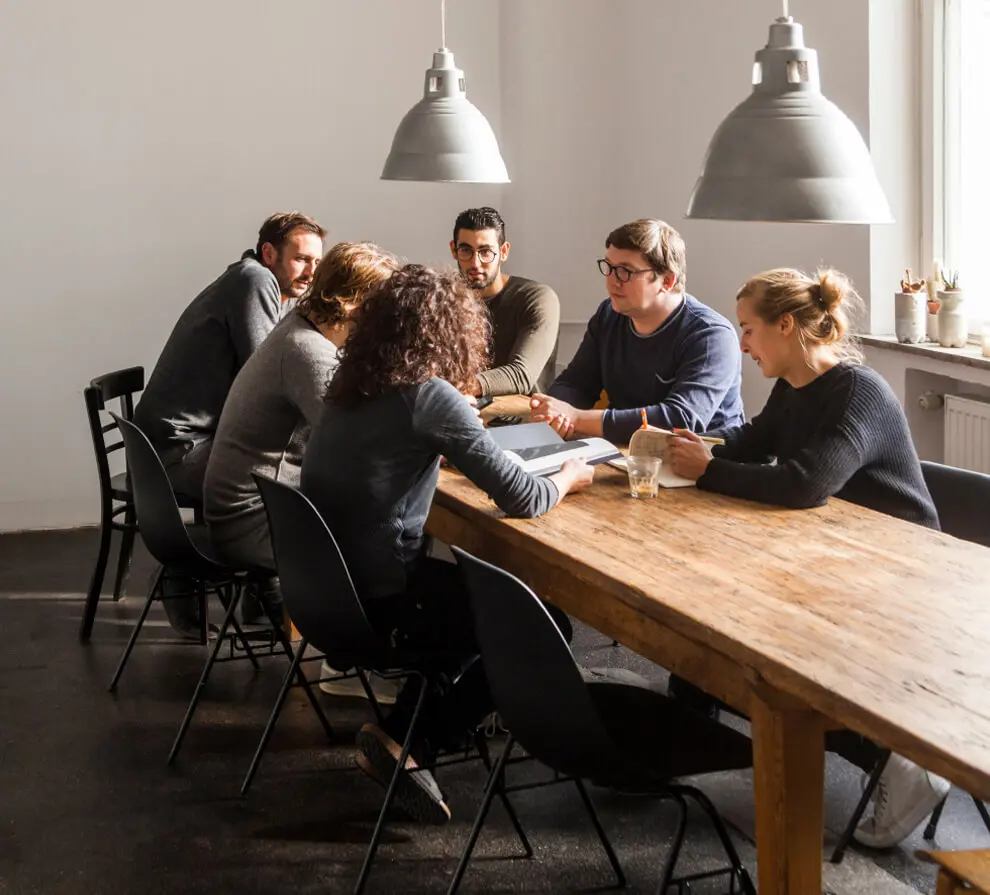 Coworkers in a modern office gather at a long wooden table, talking and flipping through books