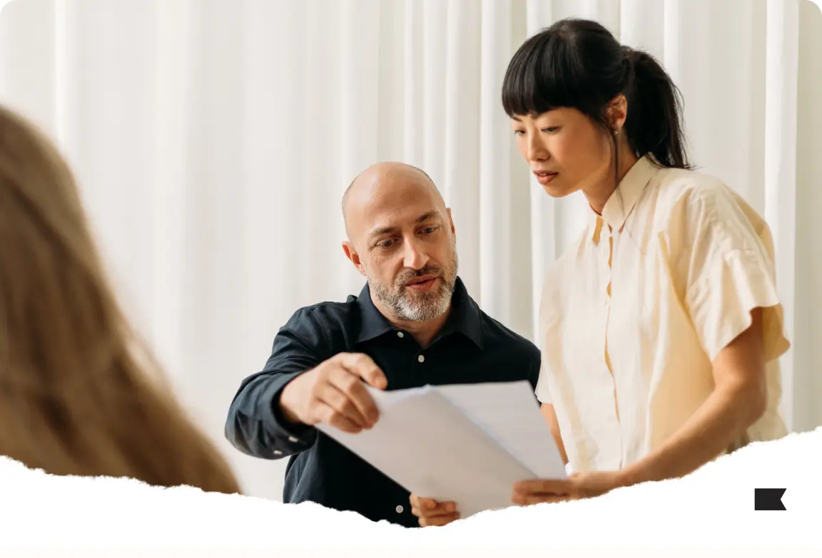 Two people reviewing a document together, one pointing at the paper. A third person is partially visible in the foreground.
