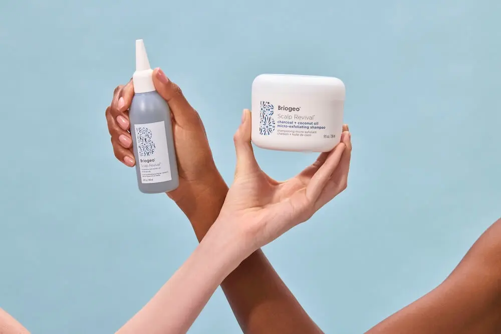 Hands holding Briogeo Scalp Revival hair products against a light blue background.