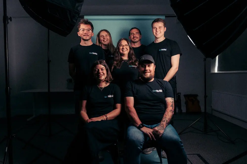 A group of seven people wearing "Get Better" branded shirts, posing together in a studio setting.