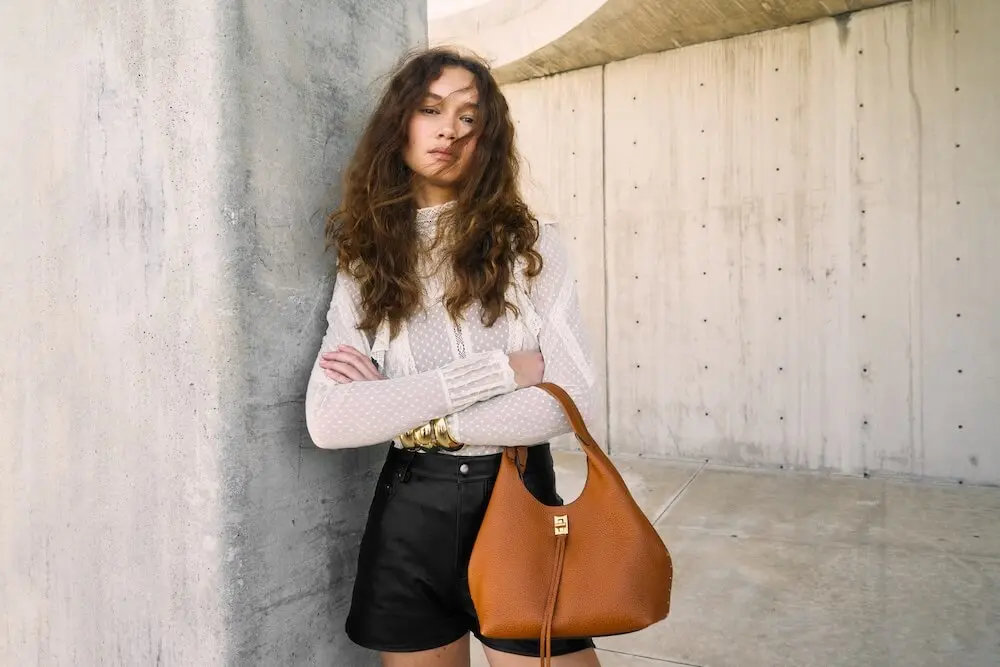 Woman with long hair in a white blouse and black shorts, holding a brown handbag, stands against a concrete wall.
