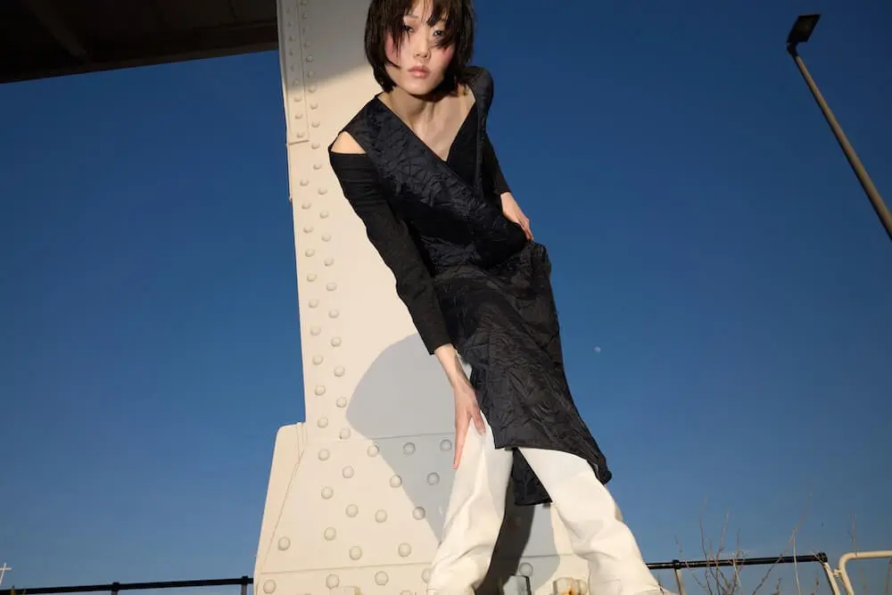 A person in a black dress and white pants poses outdoors against a metal structure, with a clear blue sky in the background.
