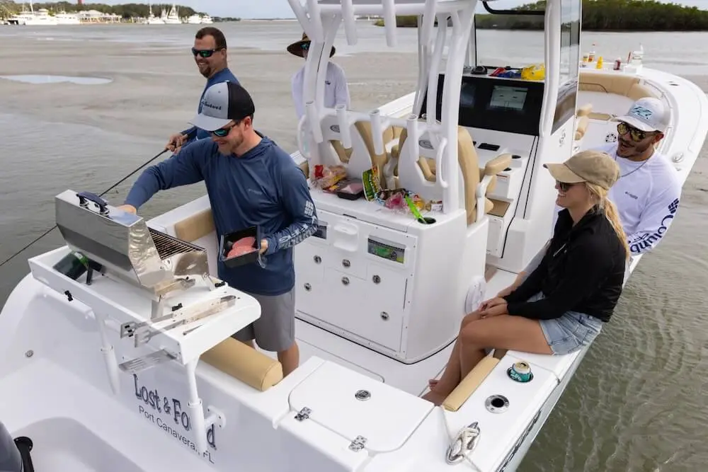 People on a boat, one grilling food, others sitting and standing, with a scenic water view.