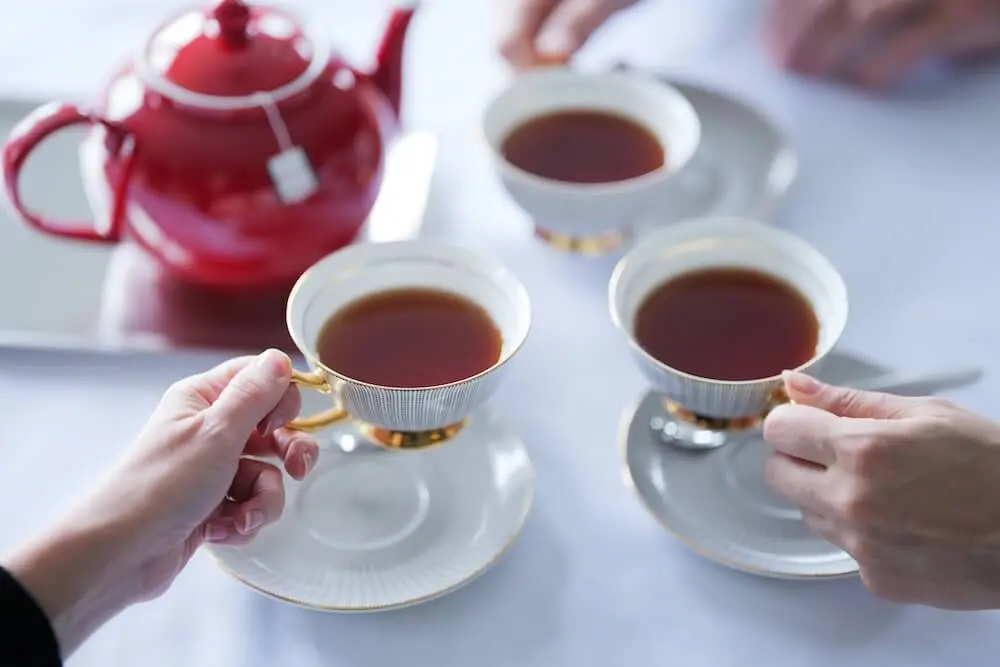 Three hands holding teacups filled with tea, with a red teapot on a tray in the background.