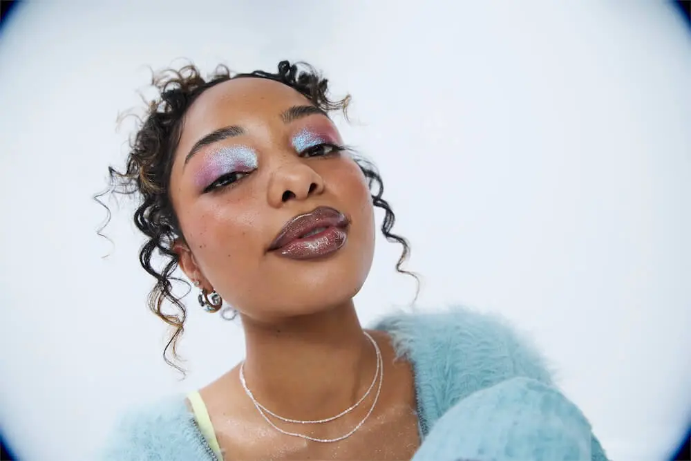 Woman with curly hair and colorful eye makeup, wearing a fluffy blue sweater and layered necklaces, looking at the camera.