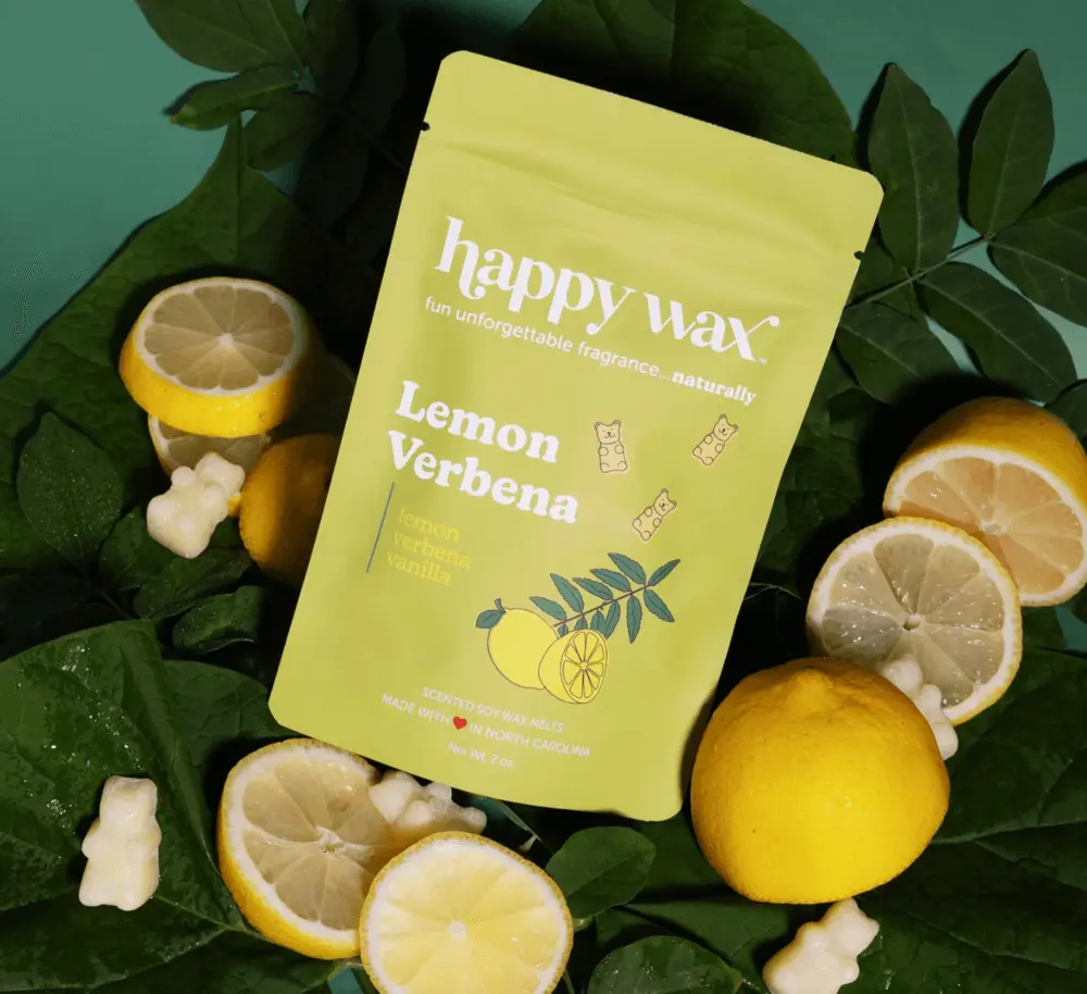 Image shows a small yellow package of Happy Wax’s flame-free scent product in Lemon Verbena on a background of leaves and surrounded by half-cut lemons and white gummy bears.